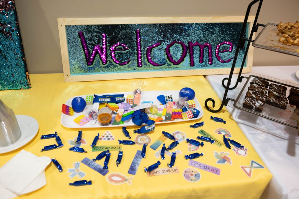 A table displays a Welcome sign with blue and pink sequins, trays of brownies and pastries, stickers, blue wrapped candies, and small inspirational items on a yellow tablecloth.