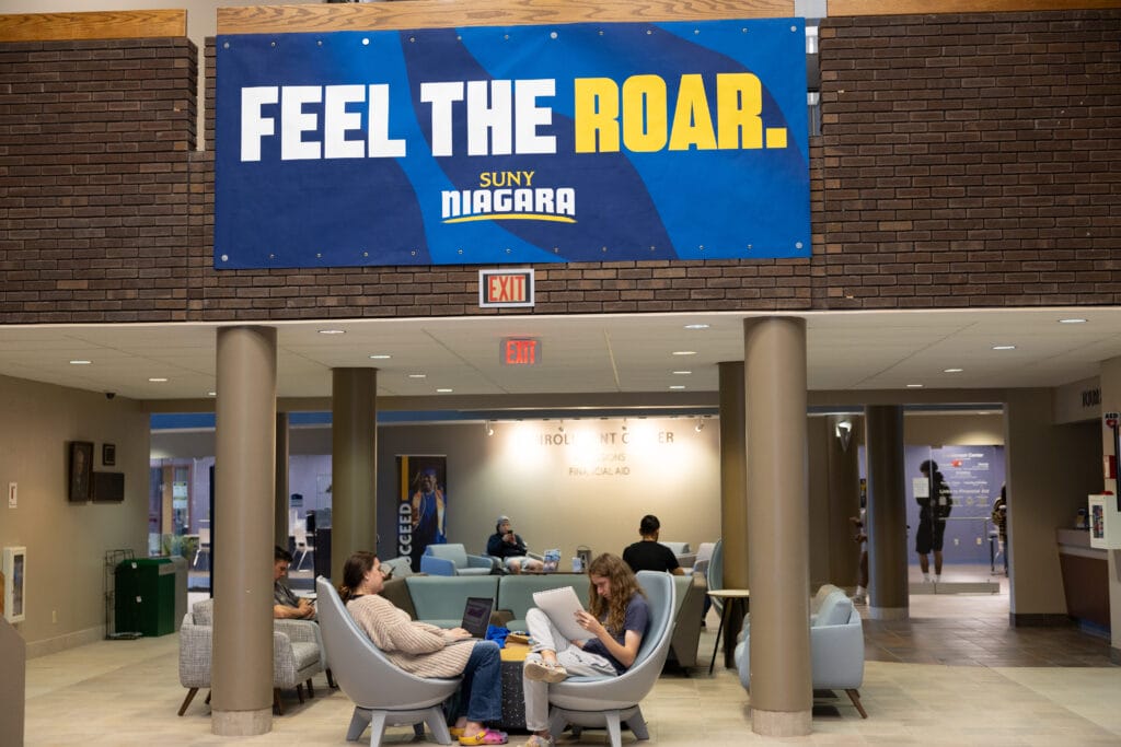 Students sit and study in a lounge area under a blue and yellow banner that reads FEEL THE ROAR. SUNY NIAGARA in a campus building with brick walls and modern furniture.