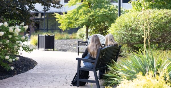 Two people with long blonde hair sit on benches along a paved garden path, surrounded by lush green trees and shrubs on a sunny day.