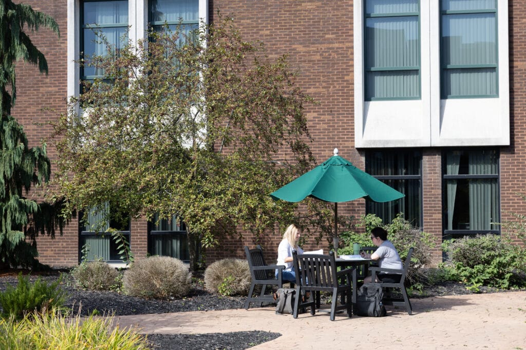 Two people sit at a table with a green umbrella, working or studying outside a brick building with large windows. Backpacks are on the ground, and trees and bushes surround the area.