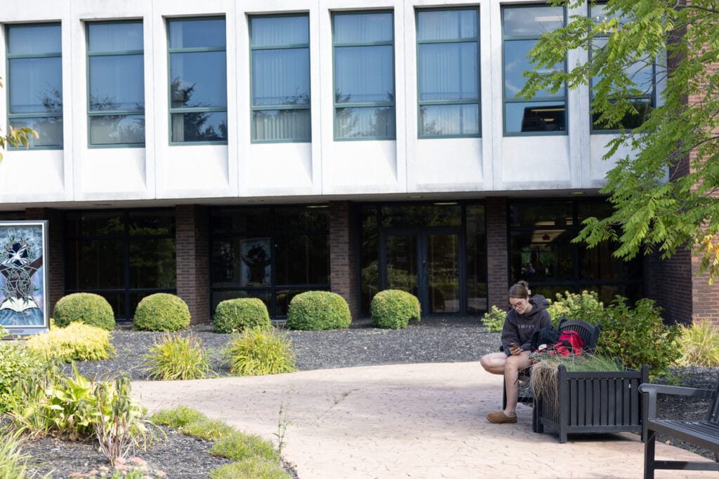 A person sits on a bench outside a modern building with large windows, looking down at a phone. There are green shrubs, plants, and trees in the landscaped area around them.