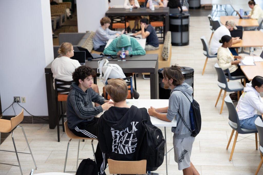Students gather in groups at tables in a bright, open study area, working on laptops, talking, and collaborating. The atmosphere is casual, with backpacks, notebooks, and various seating arrangements visible.