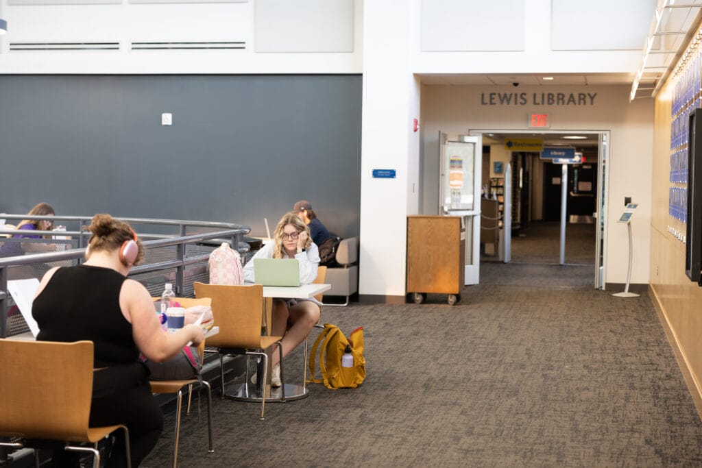 Students study at tables and use laptops near the entrance to Lewis Library. One student wears headphones, another sits with a yellow backpack and open laptop. The library entrance is in the background.