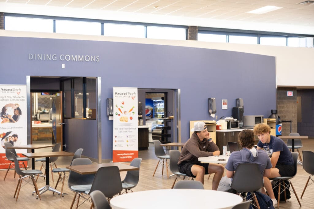Four people sit and talk at a round table in a bright dining commons area with modern decor. Nearby, empty tables, food service banners, vending machines, and beverage stations are visible.