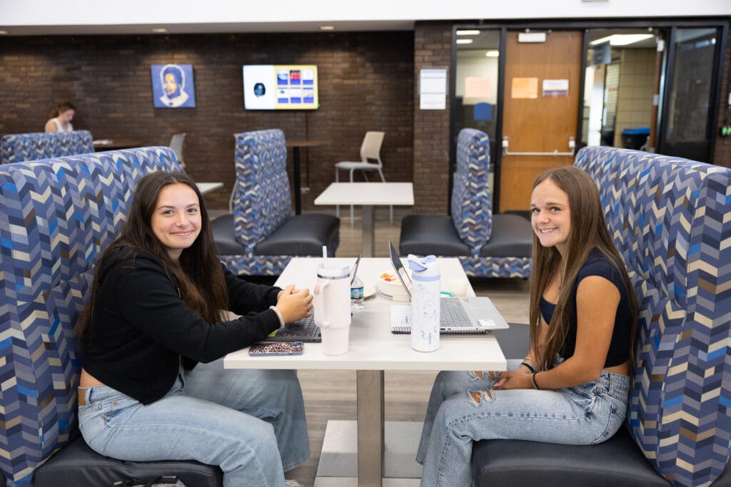 Two young women sit across from each other at a table in a lounge area with blue patterned booths, smiling at the camera. One has a laptop open and both have water bottles on the table.