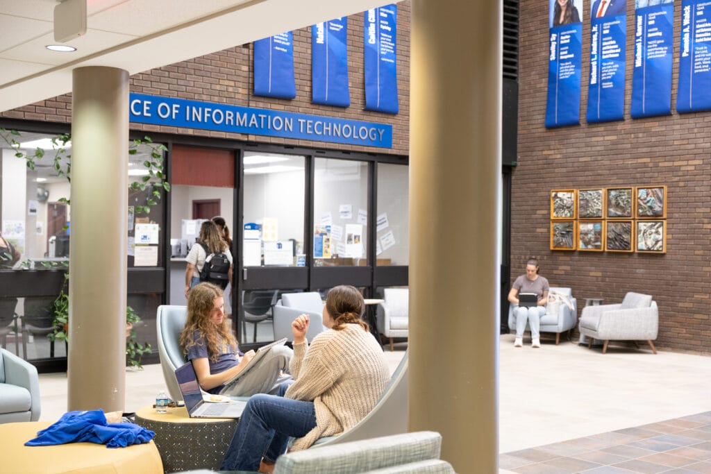 Two people sit and talk with laptops in a bright, modern lobby near the Office of Information Technology. Another person sits on a couch in the background, while someone walks into the office. Blue banners hang on the walls above.