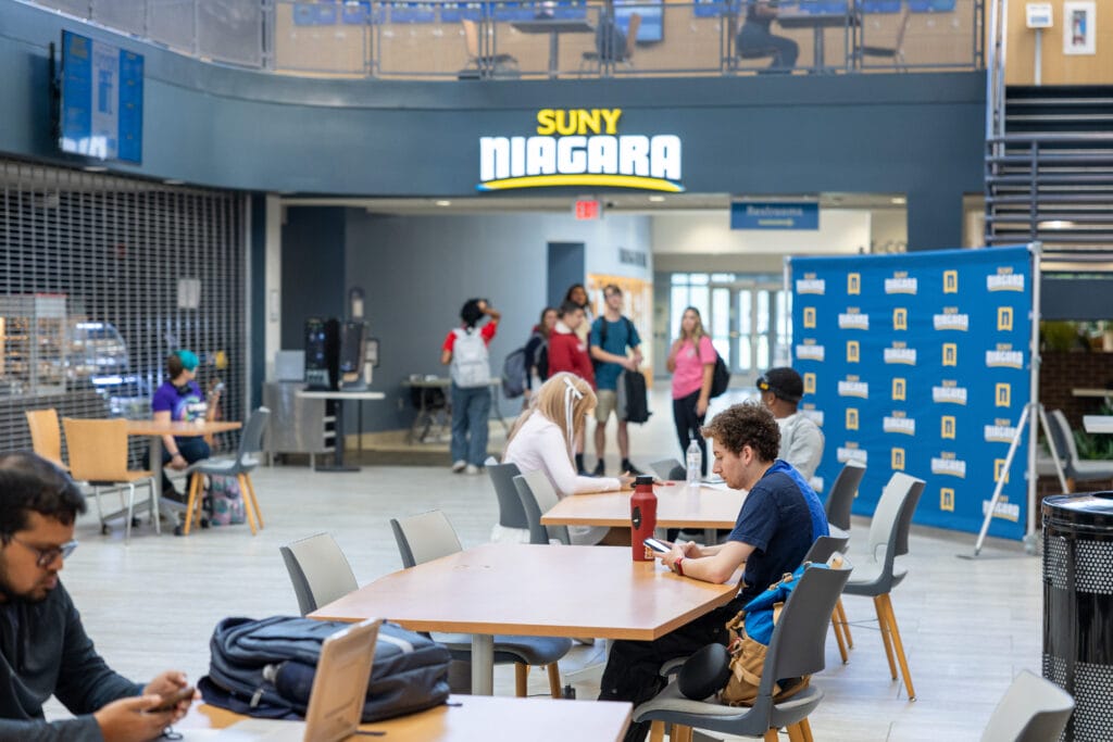 Students sit at tables and use their phones in a bright, modern common area at SUNY Niagara, while others gather and talk near the entrance under a large “SUNY Niagara” sign.