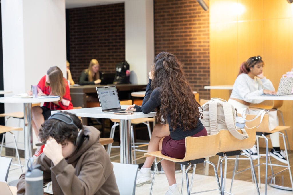 Several students work individually at tables in a brightly lit common area. Some use laptops, one wears headphones, and others are focused on their own activities. The background features brick and wood panel walls.
