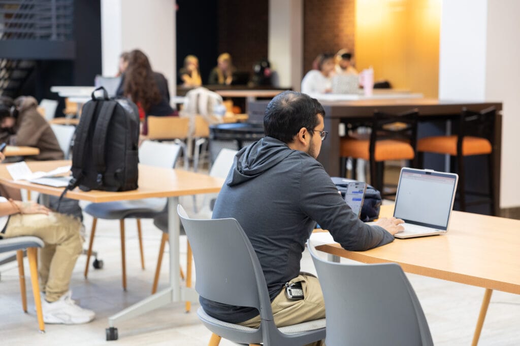 A man sits at a table using a laptop in a bright, modern study area with other people working and talking in the background. Backpacks and chairs are scattered around the tables.