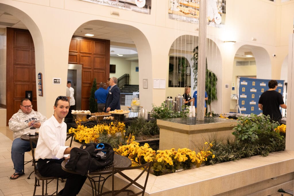 People sit at tables around a small indoor garden with yellow flowers and a water feature in a building lobby. Some eat or use phones, while others stand near a counter with food in the background.