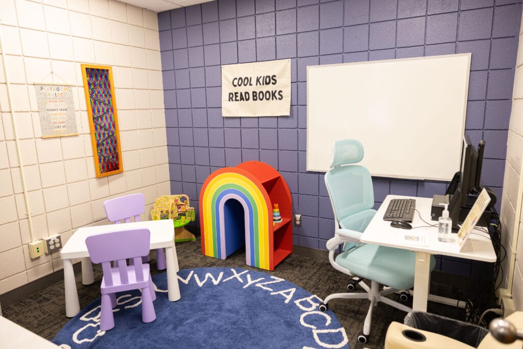 A small reading room with a purple table and two chairs, a blue rug with the alphabet, a rainbow-shaped bookshelf with books, a whiteboard, a desk with a computer, and a sign that says Cool Kids Read Books.