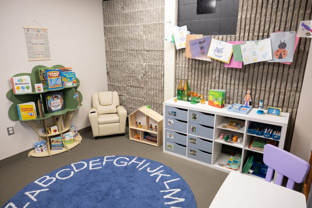 A cozy childrens reading and play corner with a recliner, bookshelf with books and games, small wooden dollhouse, dresser with toys, art on the wall, and a blue alphabet rug.