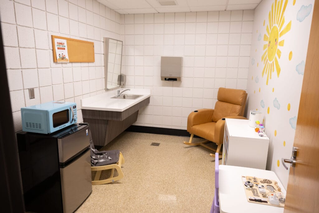 A cozy room with a tan armchair, a small refrigerator with a blue microwave on top, a sink with a mirror, children’s toys, and a wall mural featuring a sun, clouds, and birds.