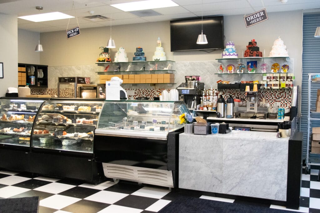 A bakery interior with black-and-white checkered floors, display cases filled with pastries, a marble counter, coffee machines, colorful decorated cakes on shelves, and signs that read ORDER HERE and PICK-UP HERE.