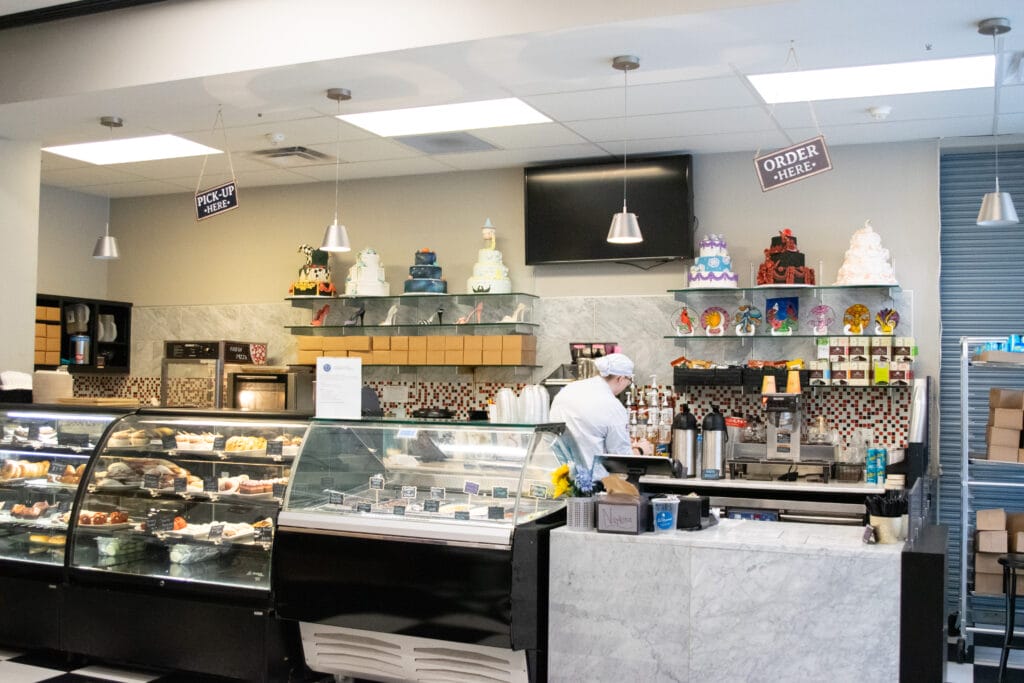 A bakery counter with display cases of pastries and cakes, shelves with colorful decorated cakes on top, and a staff member working behind the counter. Signs say Pick Up and Order Here.