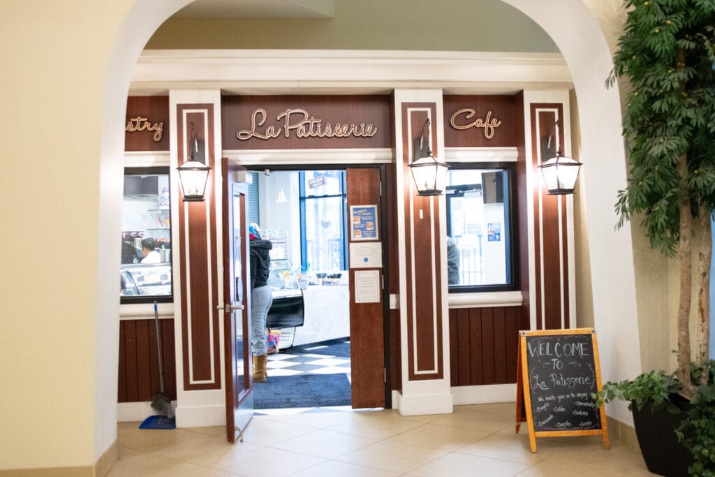 Entrance to a bakery café named La Patisserie with wooden double doors, hanging lantern lights, a chalkboard welcome sign, and indoor plants. Inside, a counter and pastry display are visible.