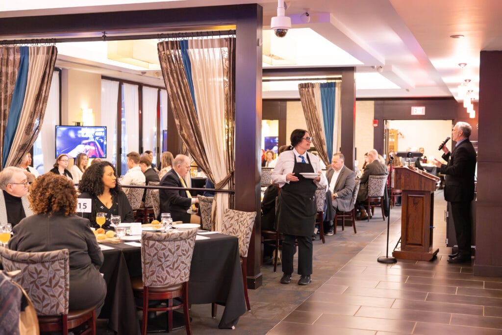 A formal dining event in a restaurant, with guests seated at tables, a waiter serving, and a man speaking at a podium. The room is well-lit and decorated, and people are engaged in conversation.