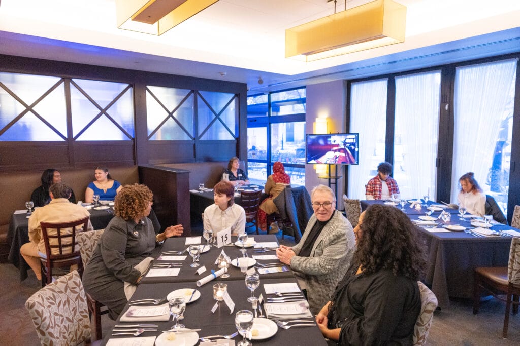 People sit at tables in a restaurant-style room set for a formal event, conversing and waiting for food. A TV screen displays a video call, and menus and glasses are arranged on the tables.