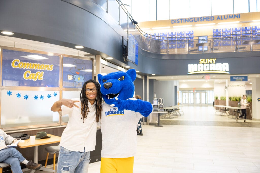 A student poses and makes a peace sign next to a blue panther mascot in a SUNY Niagara lobby near the Commons Café. Bright indoor setting with large windows and Distinguished Alumni sign above.
