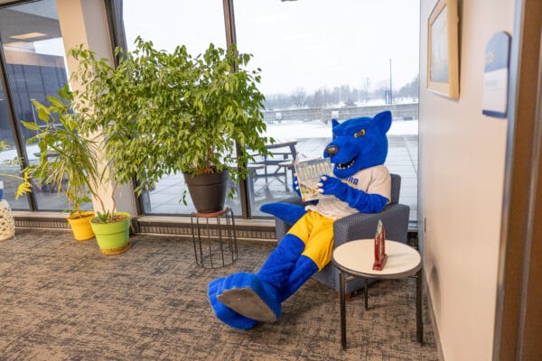 A person in a blue wolf mascot costume wearing a white shirt and yellow shorts sits in a lounge chair by a window, reading a book, with potted plants nearby and snow visible outside.