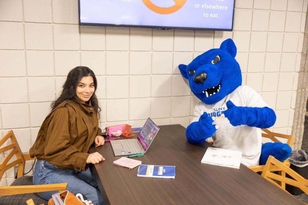 A student sits at a table with a laptop and notebook, smiling next to a person in a blue panther mascot costume giving a thumbs up. They are indoors, with a screen and books visible on the table.