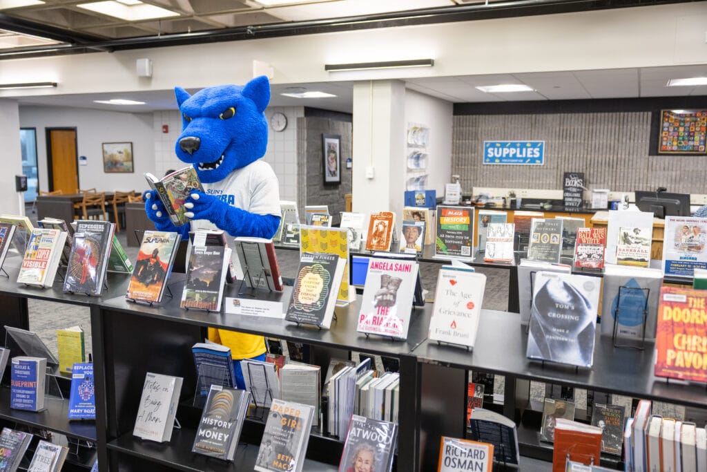 A blue panther mascot in a SUNY Cortland shirt reads a book at a table with many books on display.