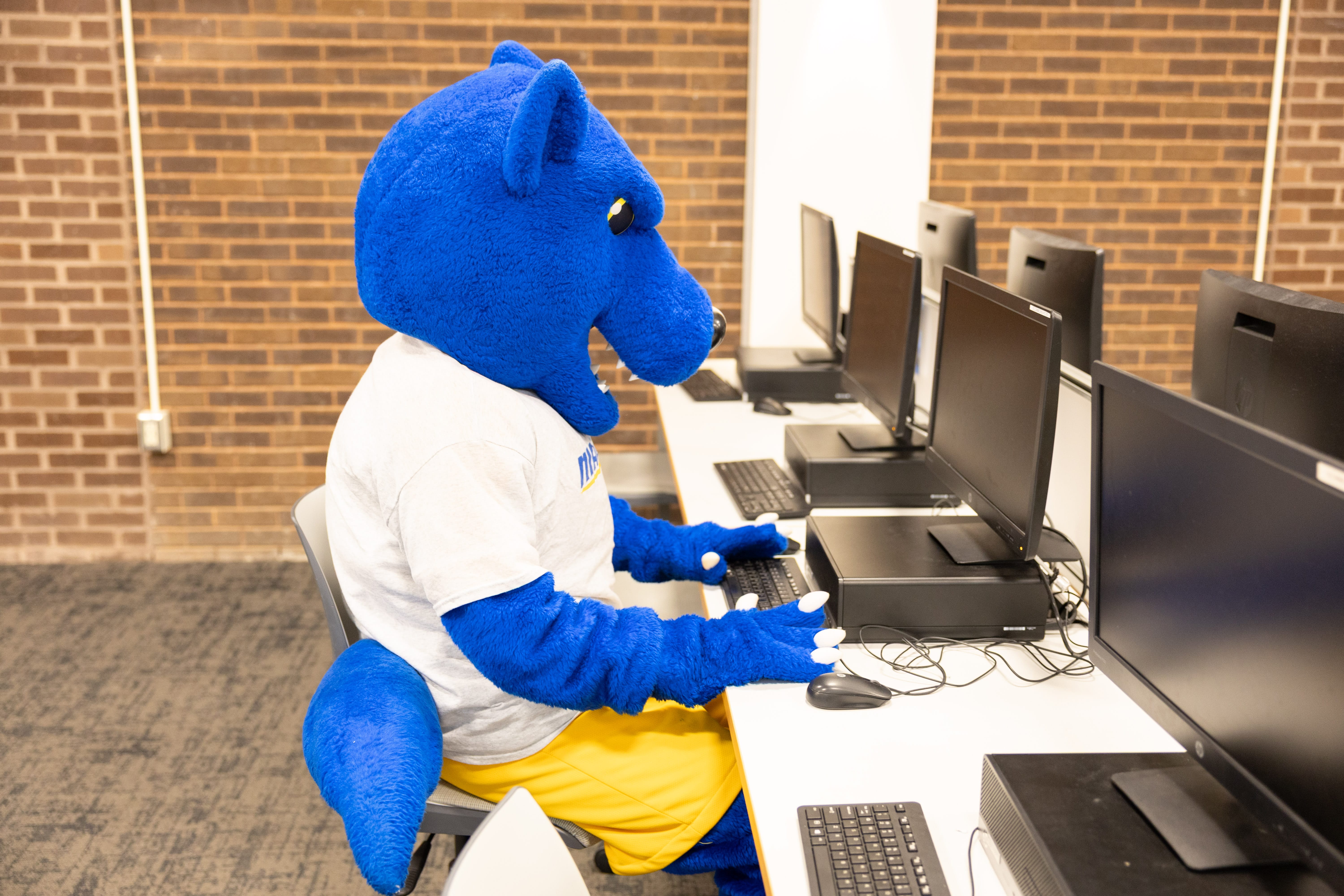 A person in a blue mascot costume sits at a row of computers in a classroom or lab, wearing a white t-shirt and yellow shorts, with hands on a keyboard.