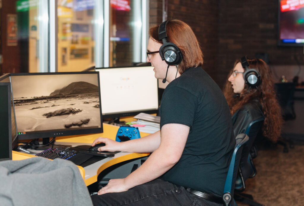 Two people wearing headsets sit at desks using desktop computers. One is typing and using a mouse, while the other looks at their screen. A blue game controller is on the desk. The setting appears to be an office or computer lab.