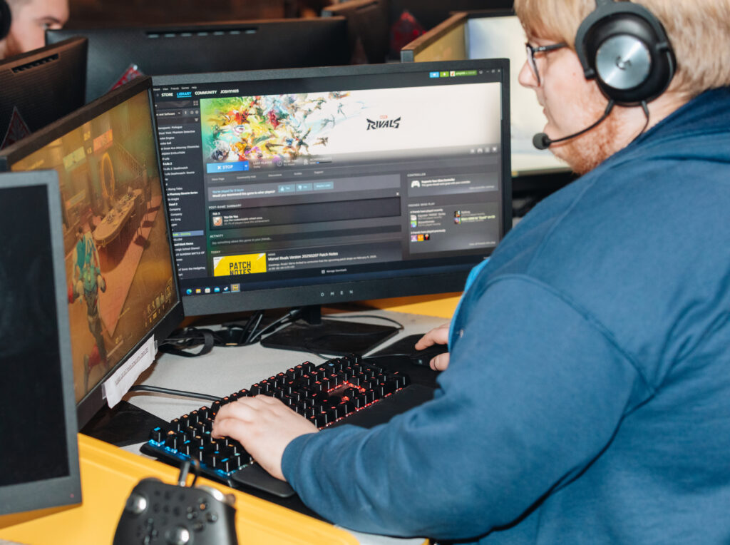 A person wearing a headset is playing a computer game at a desk with two monitors; one displays a games Steam page, and the other shows gameplay. A controller and keyboard are also visible.