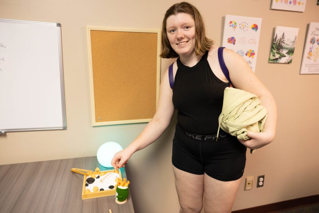 A person with short hair, wearing a black sleeveless outfit, stands indoors and smiles at the camera. They hold a light-colored garment and point at small objects on a table beside a glowing light. Artworks are on the wall behind them.