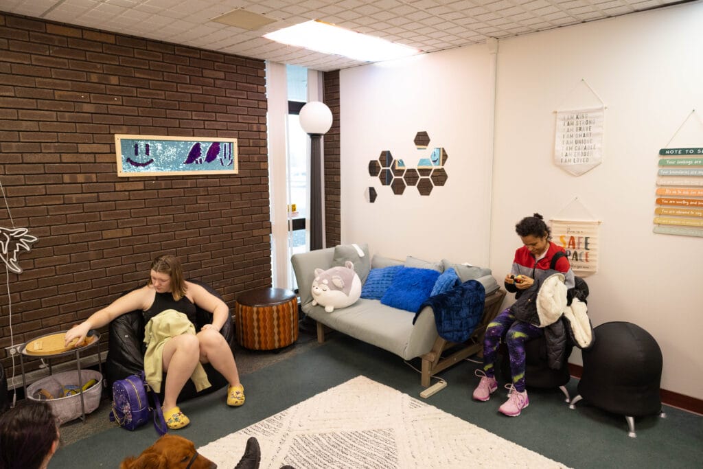 A cozy lounge with two people sitting on beanbags, one with a purple backpack, the other using a phone. The room has a gray couch, wall art, and soft lighting, creating a relaxed atmosphere.