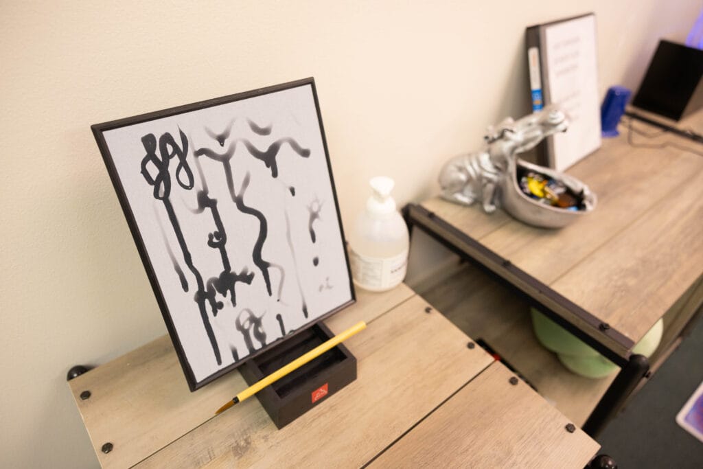 A framed abstract black and white artwork is displayed on a wooden table next to a yellow pencil and a bottle of sanitizer. In the background, there is a silver hippopotamus-shaped bowl with assorted objects inside.