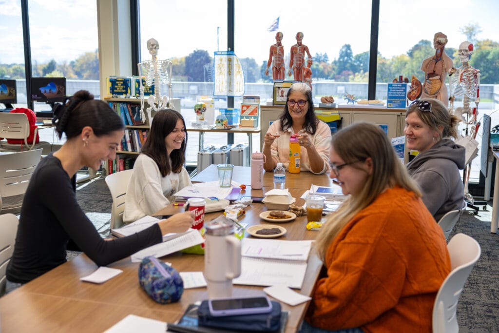 Four women sit around a table in a bright classroom or library, smiling and talking. Anatomical models and skeletons are displayed in the background. Books, snacks, drinks, and papers are spread out on the table.