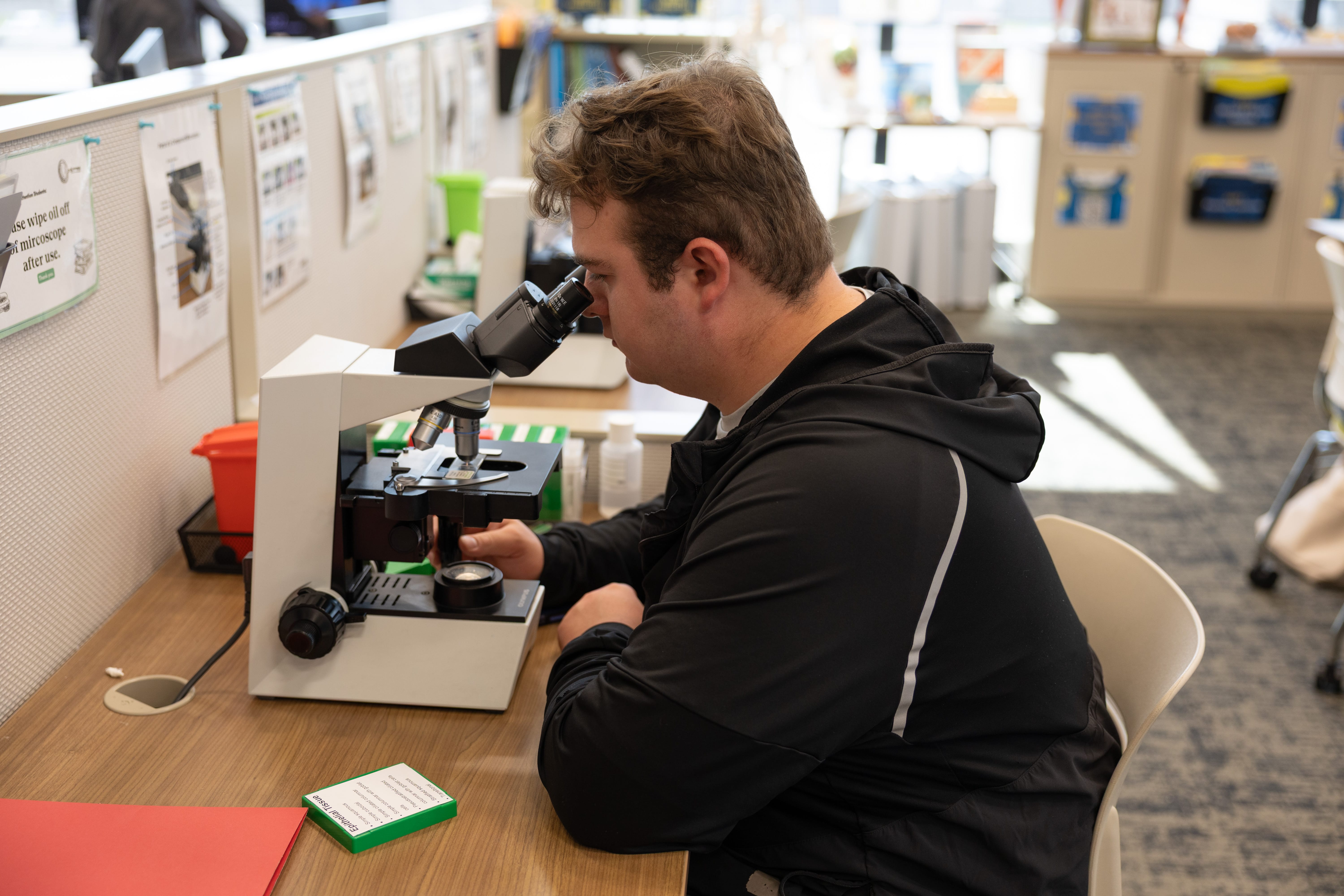 A person wearing a black jacket looks into a microscope at a desk in a brightly lit room, with educational materials and posters visible in the background.