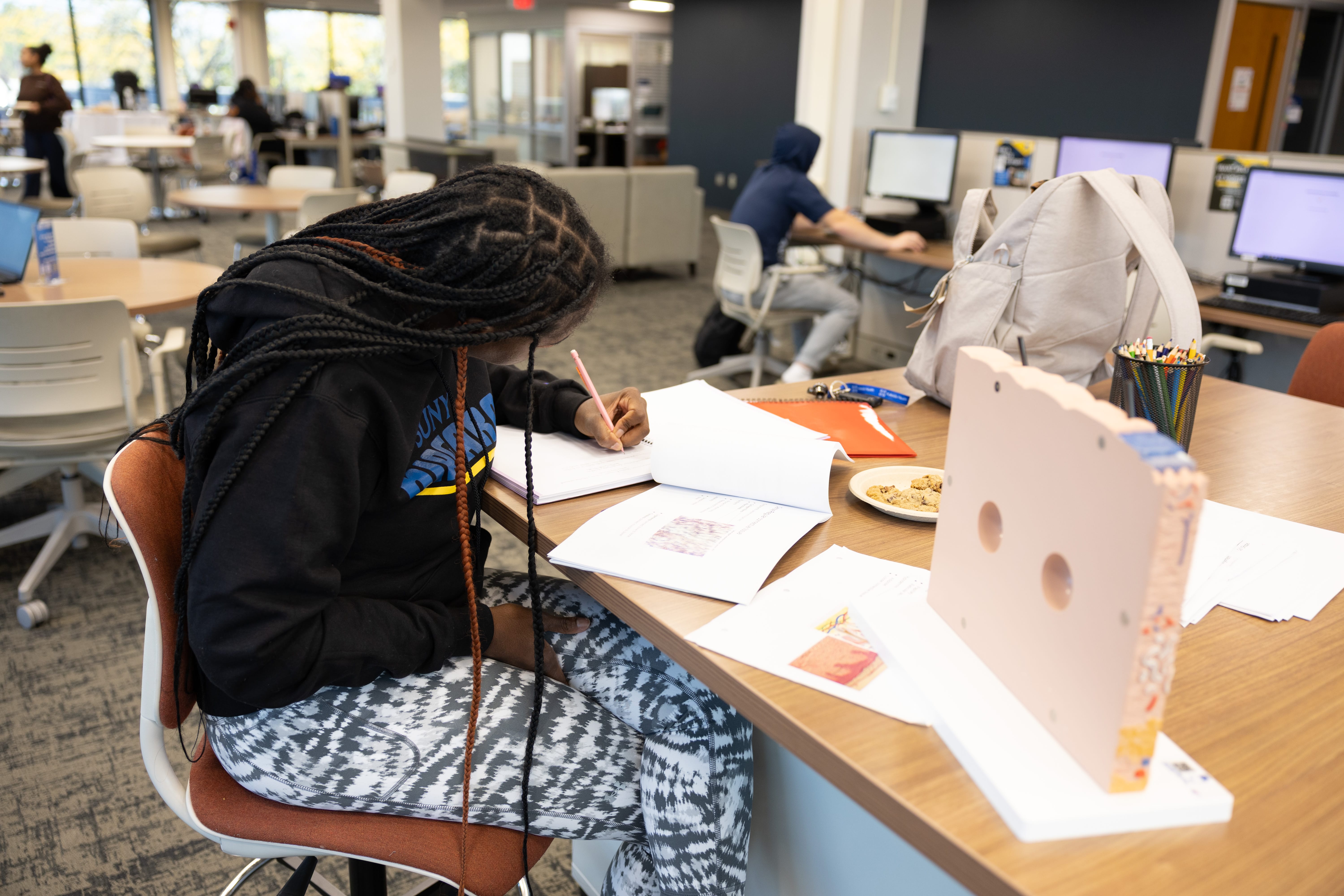 A student with long braids sits at a table in a library or study area, writing in a notebook with open books, pens, and snacks nearby. Other people are working at computers in the background.