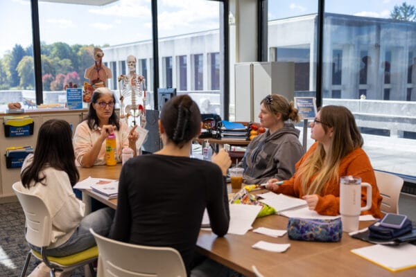 Four students sit around a table with papers and drinks, listening to an instructor in a classroom. Anatomical models and educational materials are visible, with large windows letting in natural light.