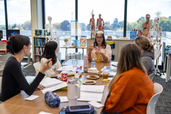A teacher explains a model of a human hand to four students seated around a table with books, papers, and snacks in a classroom filled with anatomical models and skeletons. Large windows show a bright outdoor scene.