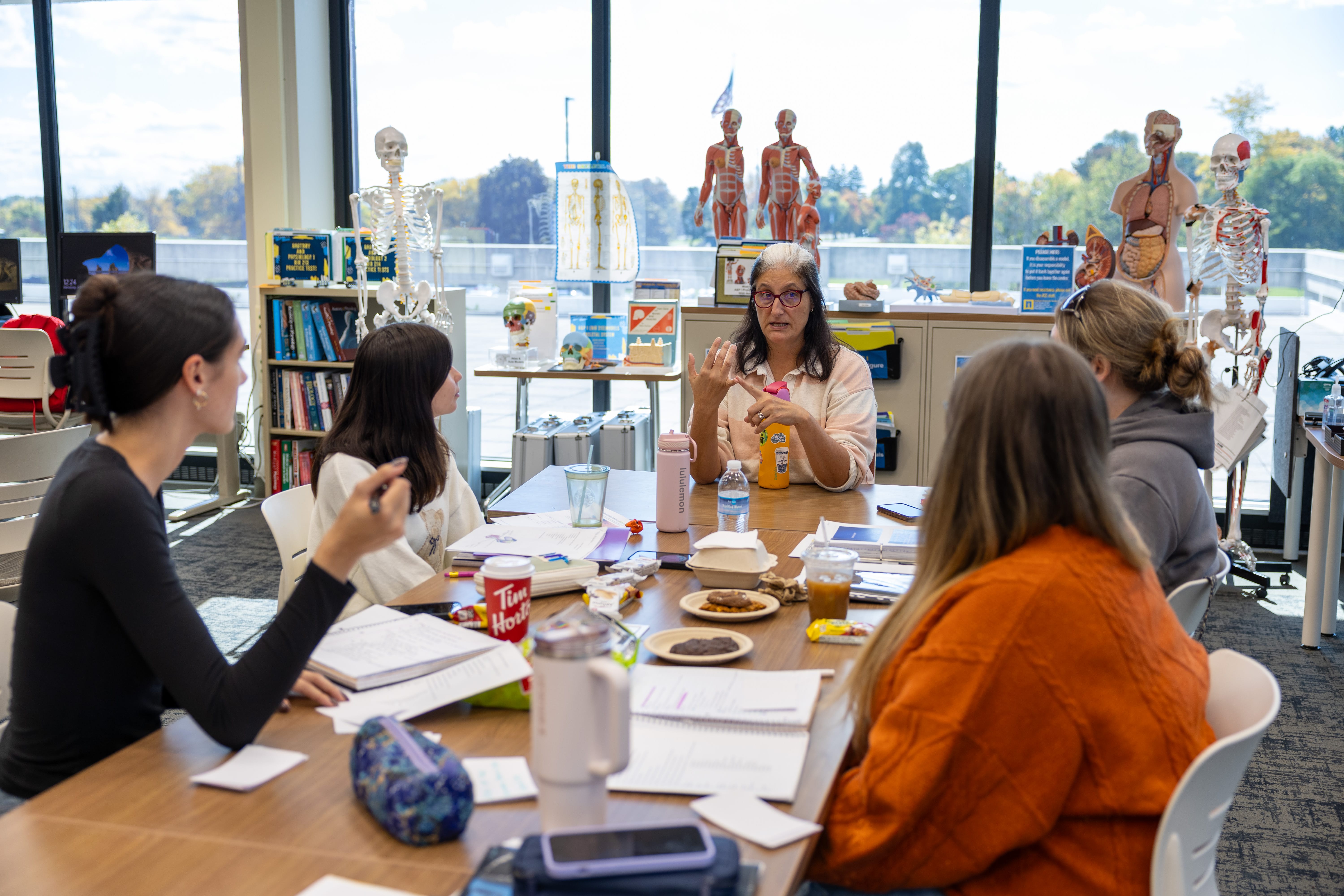 A teacher explains a model of a human hand to four students seated around a table with books, papers, and snacks in a classroom filled with anatomical models and skeletons. Large windows show a bright outdoor scene.
