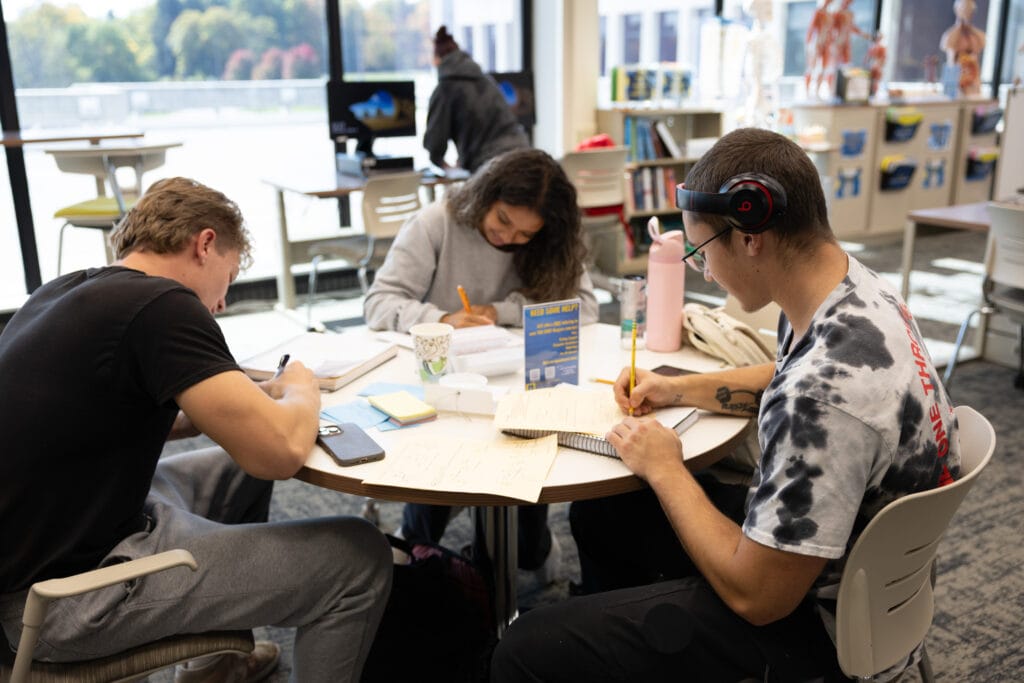 Three students sit around a table in a bright room, studying and writing in notebooks. Books, papers, and cups are on the table. Anatomical models and large windows are visible in the background.
