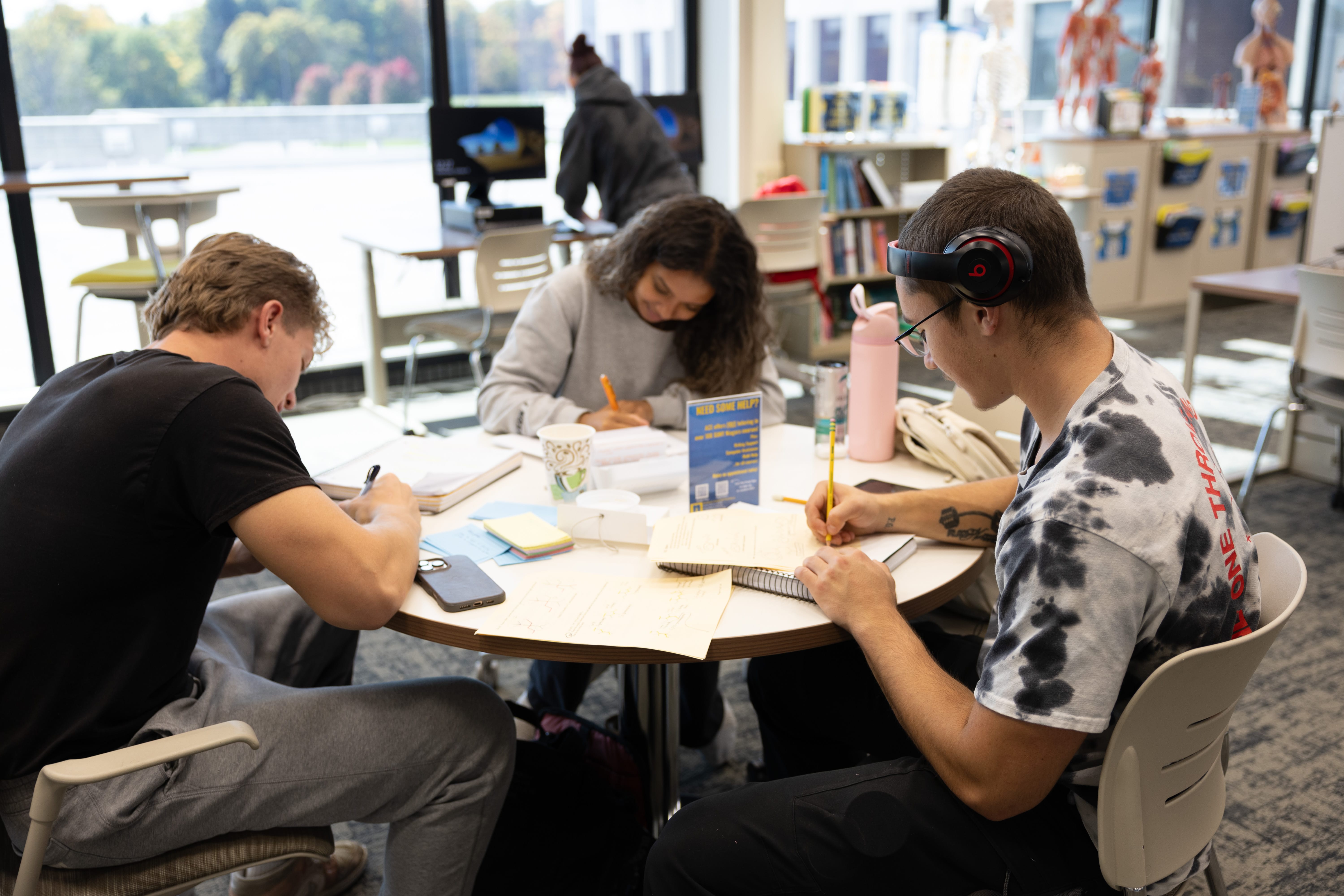 Three students sit around a table in a bright room, studying and writing in notebooks. Books, papers, and cups are on the table. Anatomical models and large windows are visible in the background.