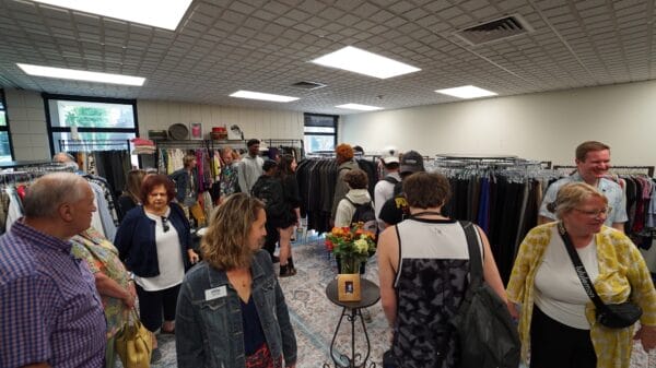 A group of people browse clothing racks and mingle in a brightly lit thrift or consignment shop, with flowers on a small table and large windows in the background.