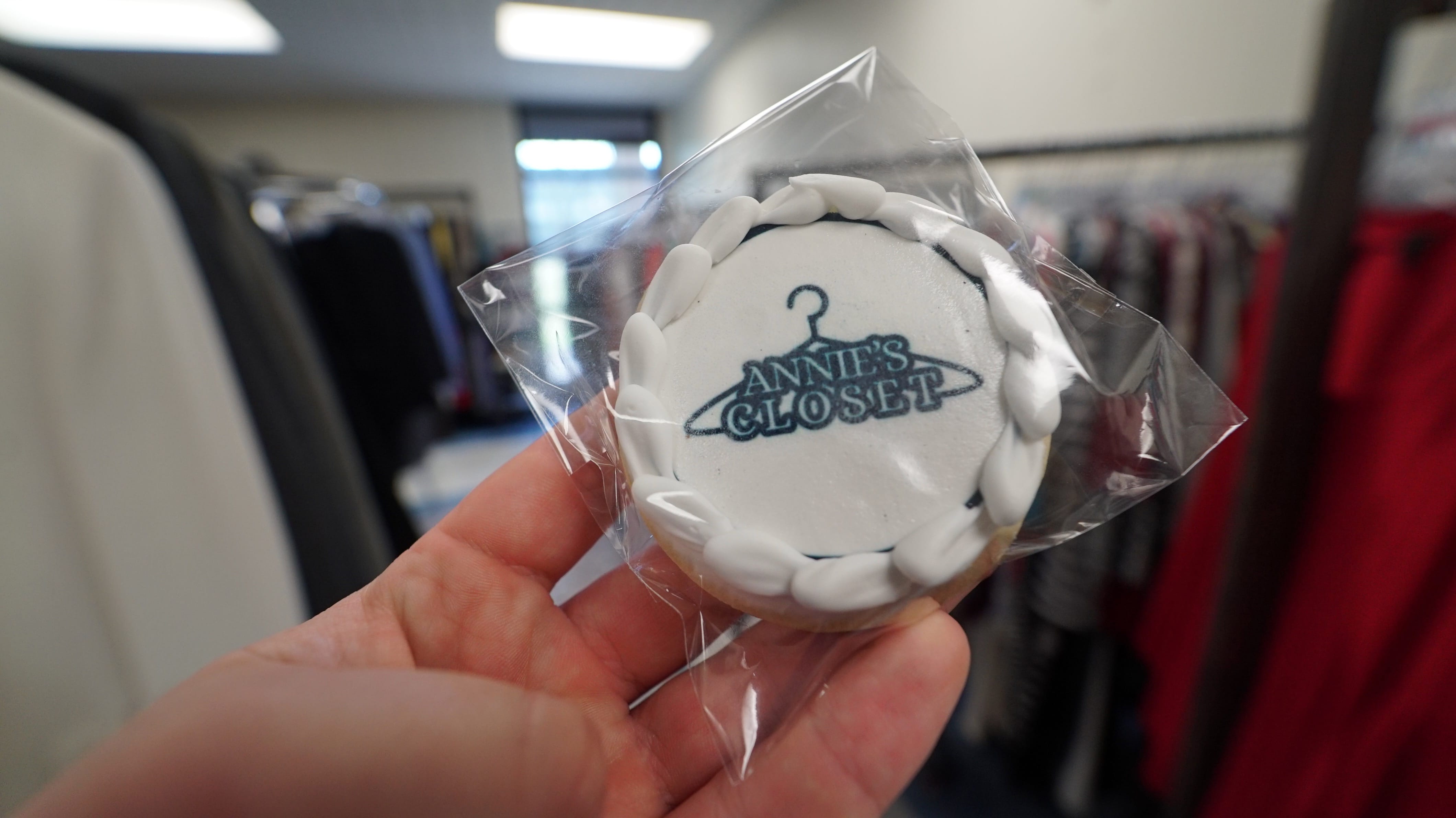 A hand holds a frosted cookie in clear plastic packaging. The cookie is decorated with a white border and features the text ANNIES CLOSET with a clothes hanger logo. Racks of clothes are blurred in the background.