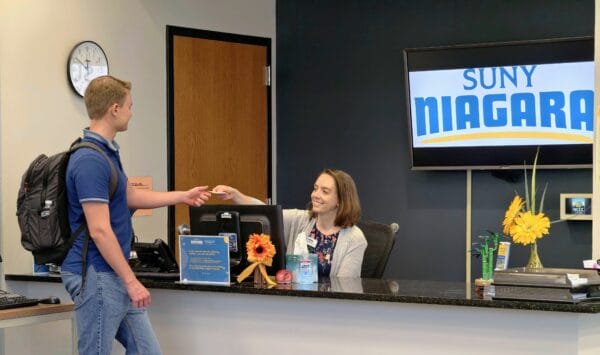 A male student with a backpack hands a card to a smiling woman at a reception desk with a SUNY Niagara sign on the wall behind her. The desk has a computer, flowers, and office items on it.