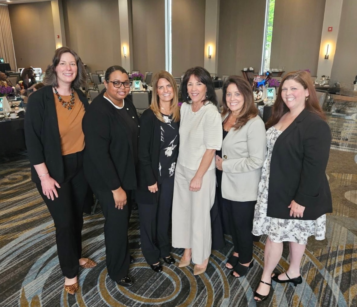 Six women stand together in a conference room, dressed in business or business-casual attire. They are smiling at the camera, and tables with items can be seen in the background.