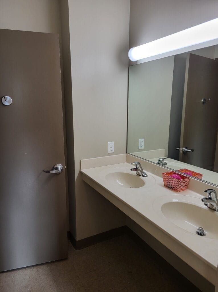 A restroom in the Student Housing Village at SUNY Niagara features two sinks, a large mirror, beige countertop, brown door partially open, and a pink basket under a bright overhead light. Apply now for the 2024-2025 academic year!.