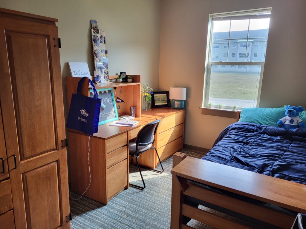 A tidy dorm room in the Student Housing Village at SUNY Niagara features a wooden bed, blue bedding, desk, chair, and wardrobe. Sunlight streams in, creating a cozy study space. Apply Now for 2024-2025 Academic Year!.