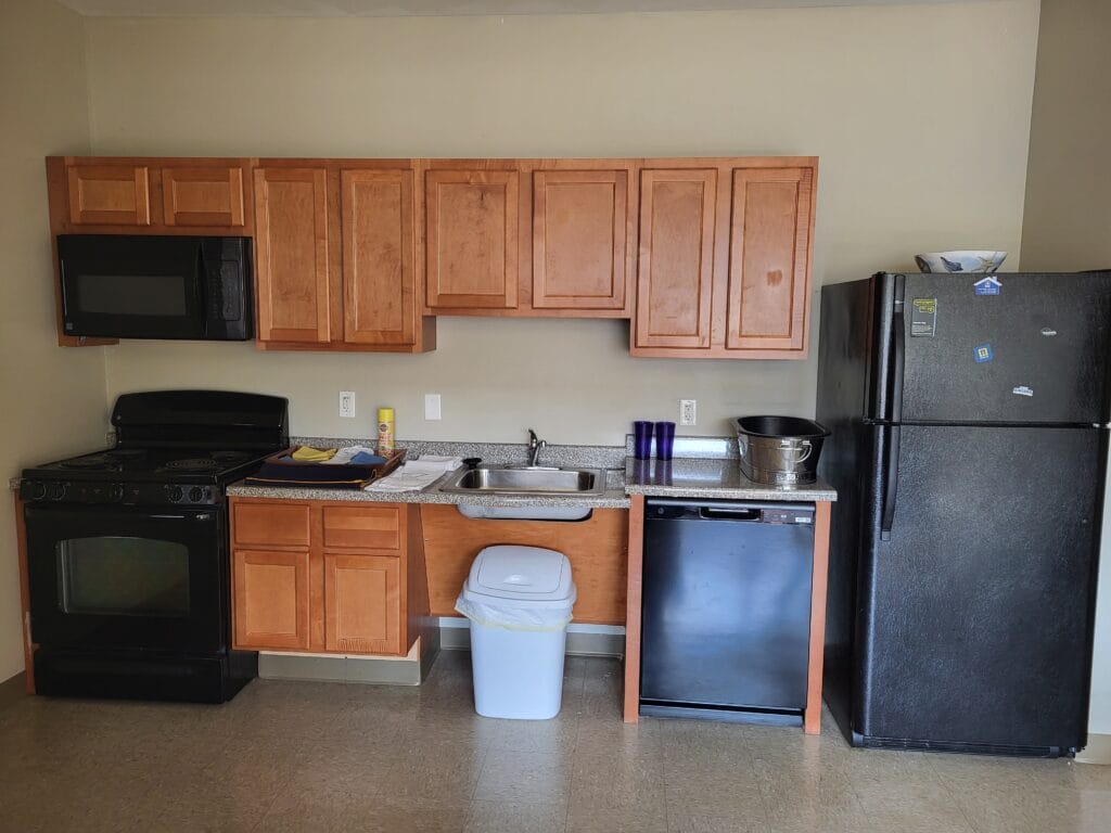 A small kitchen at Student Housing Village at SUNY Niagara features wooden cabinets, a black stove with oven, black microwave, refrigerator with magnets, dishwasher, trash can, and a sink with cups and a bowl. Apply now for 2024-2025 Academic Year!.