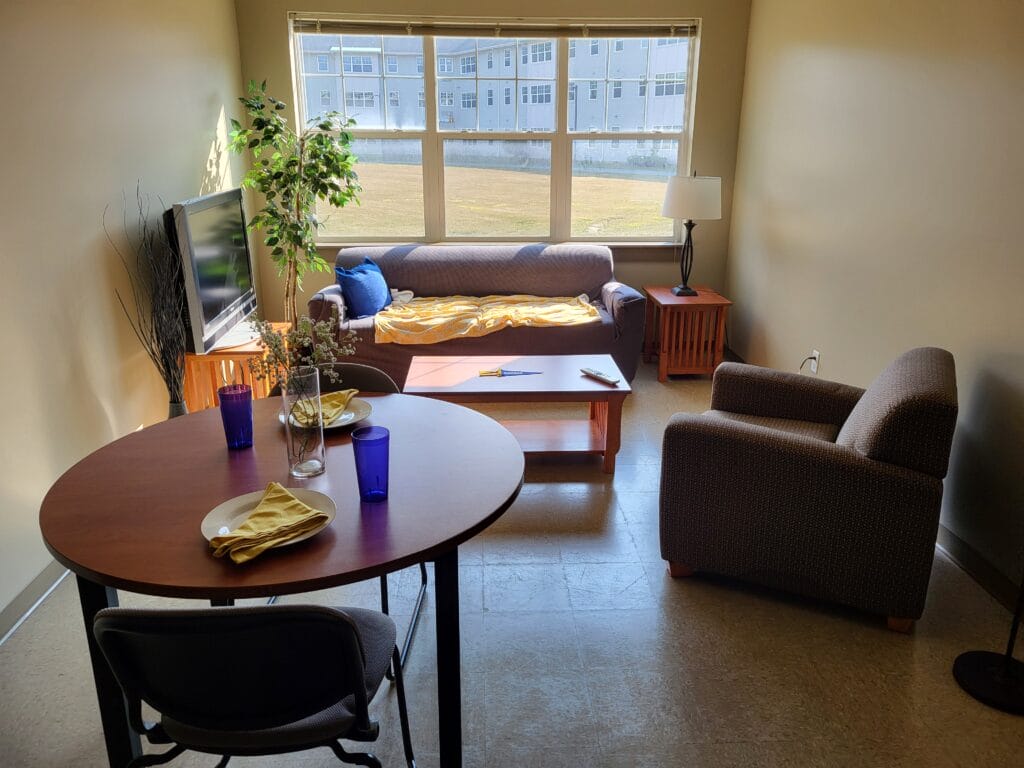 A sunlit living room in the Student Housing Village at SUNY Niagara features a sofa, armchair, TV, coffee table, and floor lamp. A dining area sits in the foreground. Apply now for the 2024-2025 academic year! Large windows let in natural light.