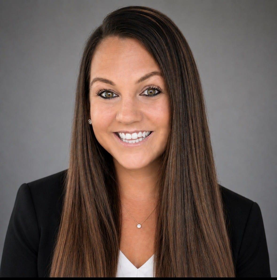 A woman with long straight brown hair, wearing a black blazer over a white top, smiles at the camera against a plain gray background.