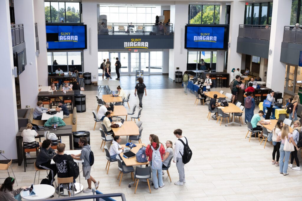Students gather, talk, and study at tables in a bright, spacious SUNY Niagara campus common area. Large windows, digital signage, and a second-floor overlook are visible. Some students walk while others sit in groups.